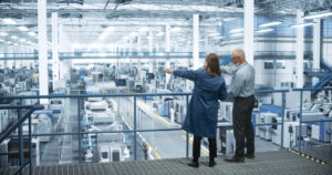 Experienced Male and Female Engineers Standing on a Platform with Their Back to Camera, Using Laptop Computer and Discussing Production at a Modern AI Automated Electronics Manufacture